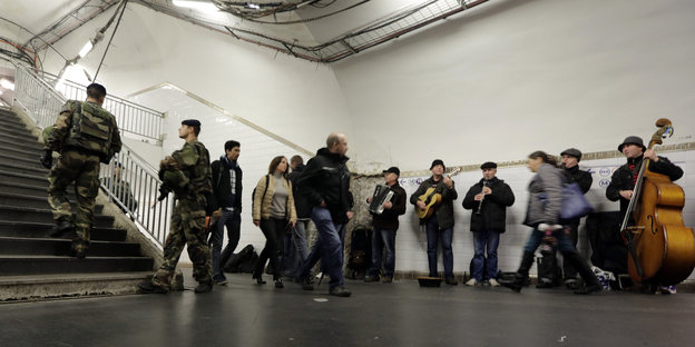 Soldaten und Passanten laufen durch einen Gang. An der Seite stehen Musiker.