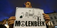 Frau hält Plakat hoch mit einer Stadtsilhouette und der Aufschrift "Molenbeek", im "o" ist ein Peace-Zeichen
