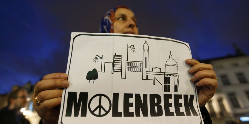 Frau hält Plakat hoch mit einer Stadtsilhouette und der Aufschrift "Molenbeek", im "o" ist ein Peace-Zeichen