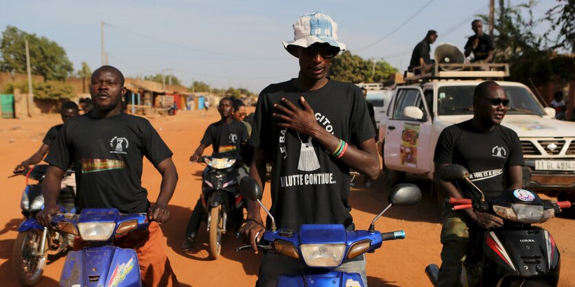 In der Bildmitte steht ein junger Mann mit Hut und Sonnenbrille. Die Hand liegt auf seiner Brust, er singt die Nationalhymne. Rechts und Links von ihm sitzen und stehen ebenfalls junge Männer. Sie alle tragen schwarze T-Shirts der Bürgerbewegung.