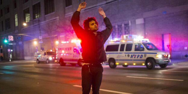 Ein Mann steht nachts auf einer Straße und hebt die Hände hoch, im Hintergrund Polizeiwagen