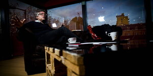 Michel Onfray sitzt mit ausgebreiteten Armen auf dem Sofa, vor sich eine Kaffeetasse, hinter sich ein Stadtpanorama