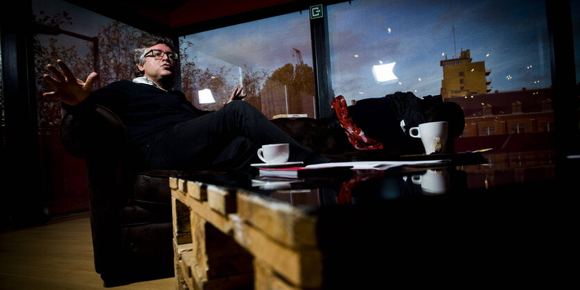 Michel Onfray sitzt mit ausgebreiteten Armen auf dem Sofa, vor sich eine Kaffeetasse, hinter sich ein Stadtpanorama