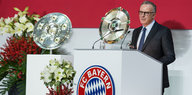 Karl-Heinz Rummenigge steht an einem Rednerpult mit FC Bayern-Logo, Trophäen und Blumen stehen daneben