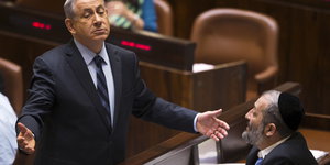 Netanjahu breitet die Arme aus und schaut nach unten. Vor ihm sitzt ein weiterer Mann mit Kippa.