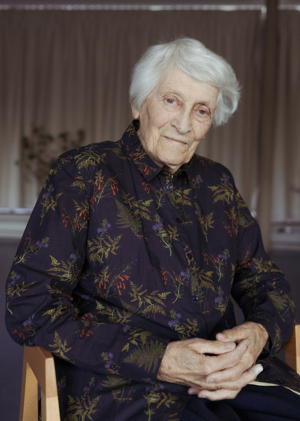 Porträt von Renate Seifert, sie sitzt auf einem Stuhl und trägt eine dunkle Bluse mit Blumenmuster