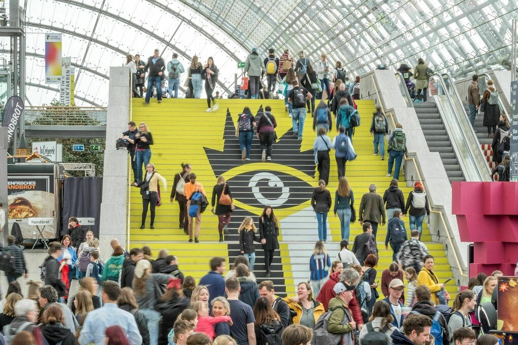 Massenandrang auf der Buchmesse, auf der gelben Treppe mit dem Logo gehen Menschen, davor staut es sich