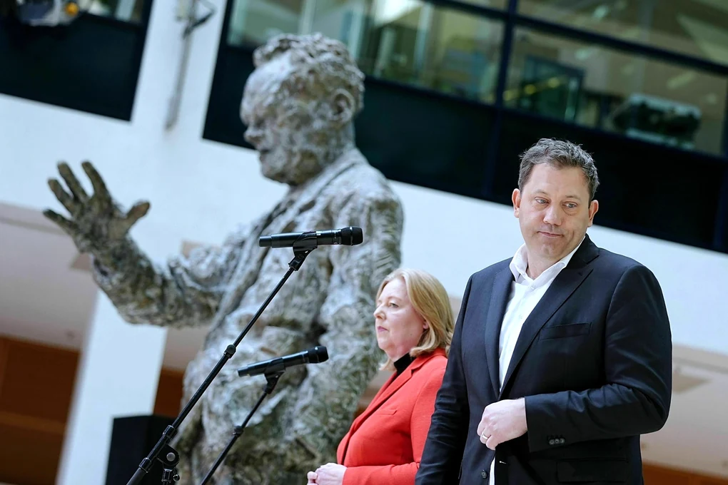 Bärbel Bas und Lars KLingbeil stehen ziemlich bedröppelt vor ihren Mikrofonen, im Hintergrund winkt die Willy-Brandt-Skulptur