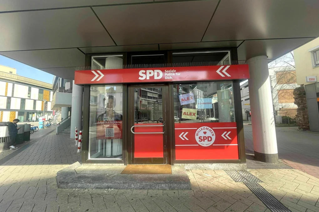 Aussenaufnahme eines SPD-Informationsbüros - eingebettet in Brutalo-Architektur - Im Fenster hängen bunte Blätter - beschriftet mit Alex, Alex. Schweizer