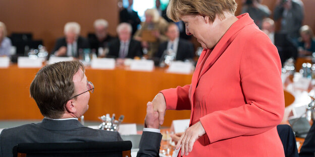 Ramelow, sitzend, und Merkel, sthehen, reichen sich die Hand