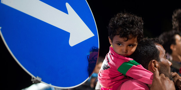 Schild mit Pfeil, der auf ein Flüchtlingskind weist, dass auf der Schulter seines Vaters sitzt.