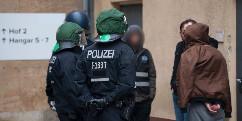 Polizisten mit Helm stehen neben Menschen in Zivil, einer hat gebundene Hände