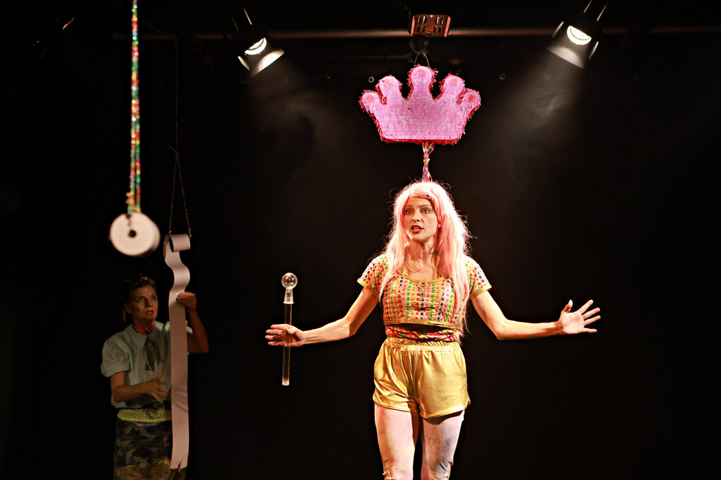 Ein Foto einer Aufführung des Theaterstücks „Pretty Privileged – Der Kaiserin neue Kleider“. Im Vordergrund ist Charlotte Alten im Bühnenlicht zu sehen, über ihr schwebt eine pinkfarbige Krone.