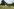 Drei Holzhütten auf einer Wiese, dahinter Bäume und Büsche