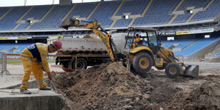 Ein Bagger und ein Bauarbeiter stehen mitten in einem unfertigen Stadion