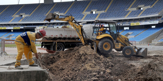 Ein Bagger und ein Bauarbeiter stehen mitten in einem unfertigen Stadion