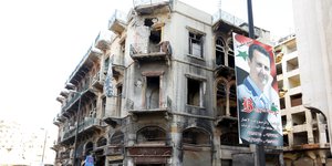 Baschar al-Assad blickt von einem Plakat herab, das an einer Straßenlaterne angebracht ist - im Hintergrund ein zerstörtes Gebäude.