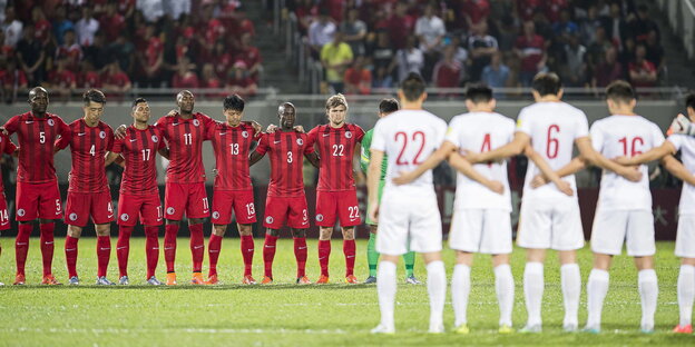 Eine Reihe Fußballspieler in roten Trikots steht mit verschränkten Armen einer Reihe Fußballspieler in weißen Trikots gegenüber