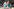 Menschen in Einhorn-Kostümen und mit Regenbogenfahnen auf einem schattigen Platz.