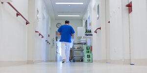 Ein Pfleger im blauen Oberteil läuft einen weißen Krankenhausflur entlang.