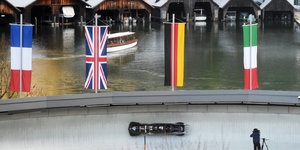 Ein Viererbob fährt auf der Kunsteisbahn am Königssee, dahinter sind Bootshütten am Seeufer zu sehen