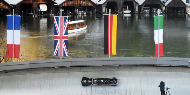 Ein Viererbob fährt auf der Kunsteisbahn am Königssee, dahinter sind Bootshütten am Seeufer zu sehen