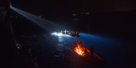 Von Scheinwerfern angestrahltes, voll besetztes Boot im Wasser bei Nacht.