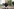 Ein Mann mittleren alters schaut skeptisch in die Ferne, hinter ihm ist eine Plattenbaulandschaft, mit Plakaten und einer Person die durchs Bild läuft