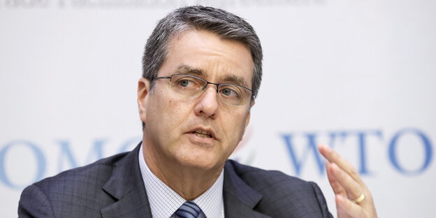 Roberto Azevedo mit Brille und Anzug sitzt vor einer Wand, auf der WTO steht