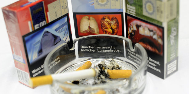 Drei Zigarettenschachteln mit Warnhinweisen und Warnfotos, davor ein Aschenbecher