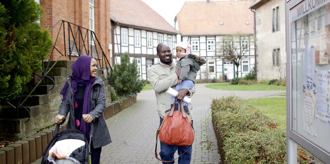 Eine Frau schiebt einen leeren Kinderwagen, ein Mann hat ein Kind auf dem Arme, im Hintergrund Fachwerkhäuser