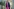 Ilse Aigner trägt ein Kleid in knalligem Pink, einen blauen Blazer, sie lächelt hat halblange dunkle Haare und eine Brille - unter dem Arm ein Labtop