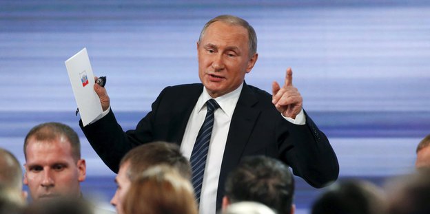 Wladimir Putin hebt bei einer Rede den linken Zeigefinger und die rechte Hand hoch
