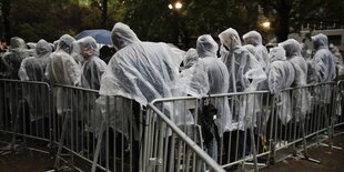 Menschen warten in Regencapes draußen hinter einem Absperrgitter.