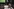 Zwei weiblich gelesene Personen, von hinten gesehen, das Gesicht der einen Person leicht angeschnitten, blicken einander an, den Arm lässig umeinandergelegt, im Hintergrund ist ein Fußballspiel zu sehen