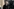 Porträt von Hermann Höllenreiner, er hat graue Haare, trägt einen dunklen Mantel und hat einen sehr ernsten Gesichtsausdruck. Im Hintergrund sind unscharf Gebäude des KZ Auschwitz-Birkenau zu sehen