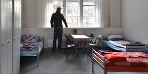 Ein Mann steht in einer Flüchtlingsunterkunft und schaut aus dem Fenster. Im Zimmer stehen drei Betten.