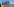 Blauer Himmel, im Vordergrund Stacheldraht, dahinter ein Mäuerchen und Menschen in Militärkleidung mit Militärlaster