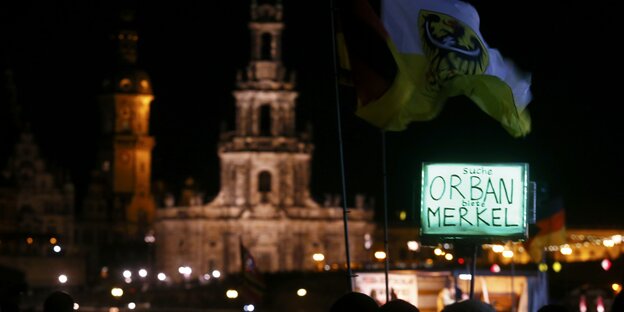 Pegida-Anhänger im nächtlichen Dresden, im Vordergrund eine blaues Schild: „Suche Orban, biete Merkel“