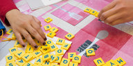 Hände schieben gelbe Plättchen mit Buchstaben über eine rot-weiße Tischdecke