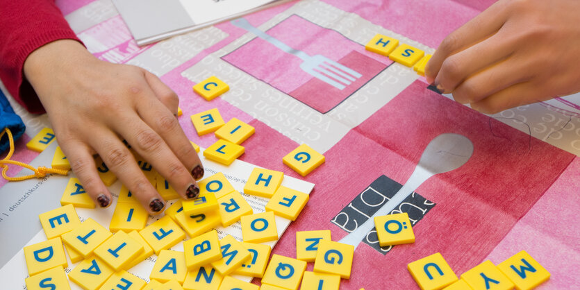 Hände schieben gelbe Plättchen mit Buchstaben über eine rot-weiße Tischdecke