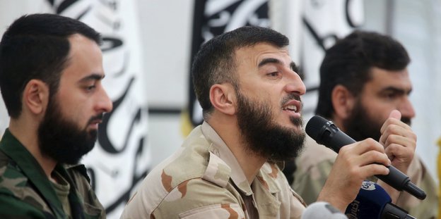 Rebellenchef Sahran Allush sitzt zwischen zwei Männer und spricht in ein Mikrofon