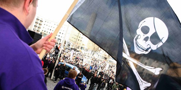 Ein Mensch im violetten Pullover hält eine schwarze Fahne mit einem weißen Totenkopf, Augenklappe und gekreuzten Knochen hoch