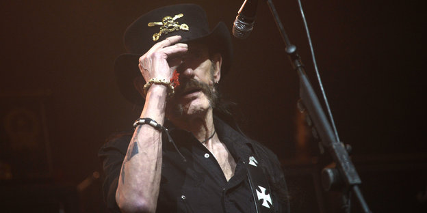 Lemmy Kilmister steht hinter einem Mikro und hält die Hand an seinen Hut