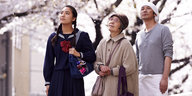 Eine junge Frau, eine ältere Frau und ein Mann mittleren Alters stehen vor blühenden Kirschbäumen und blicken nach oben