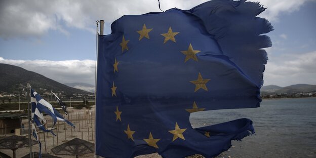 Ruinierte EU und griechische Flaggen fliegen in Fetzen von einem Fahnenmast auf einem Strand.