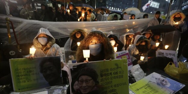 Menschen sitzen auf dem Boden mit Kerzen in der Hand und Schildern, auf denen alte Frauen zu sehen sind