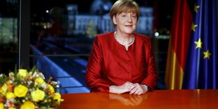 Angela Merkel steht in einem roten Blazer an einem Tisch, auf ihm stehen gelbe Blumen