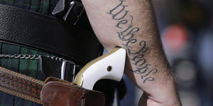 Ein Revolver in einem Holster, der Mann, der ihn trägt, hat "we, the people“ auf den Arm tätowiert