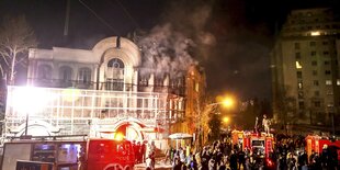 Nacht, erleuchtetes Gebäude, aus dessen Fenster Rauch steigt, davor Menschen und ein Feuerwehrauto